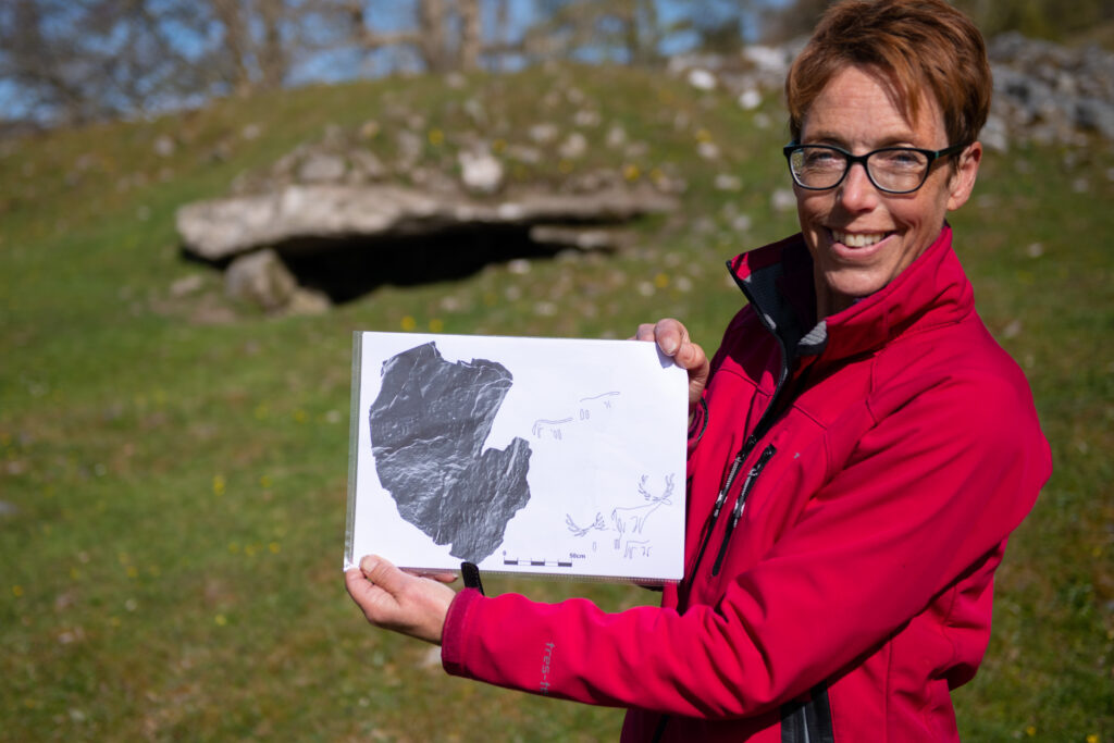 Photo of a woman in a red jacket and glasses holding a drawing of deer in front of a cairn