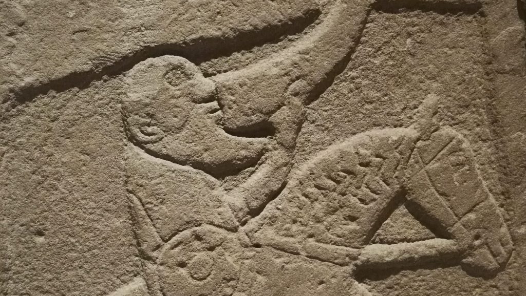 Photo of a carved stone featuring an old man with pointed beard riding an old ass and drinking from a horn