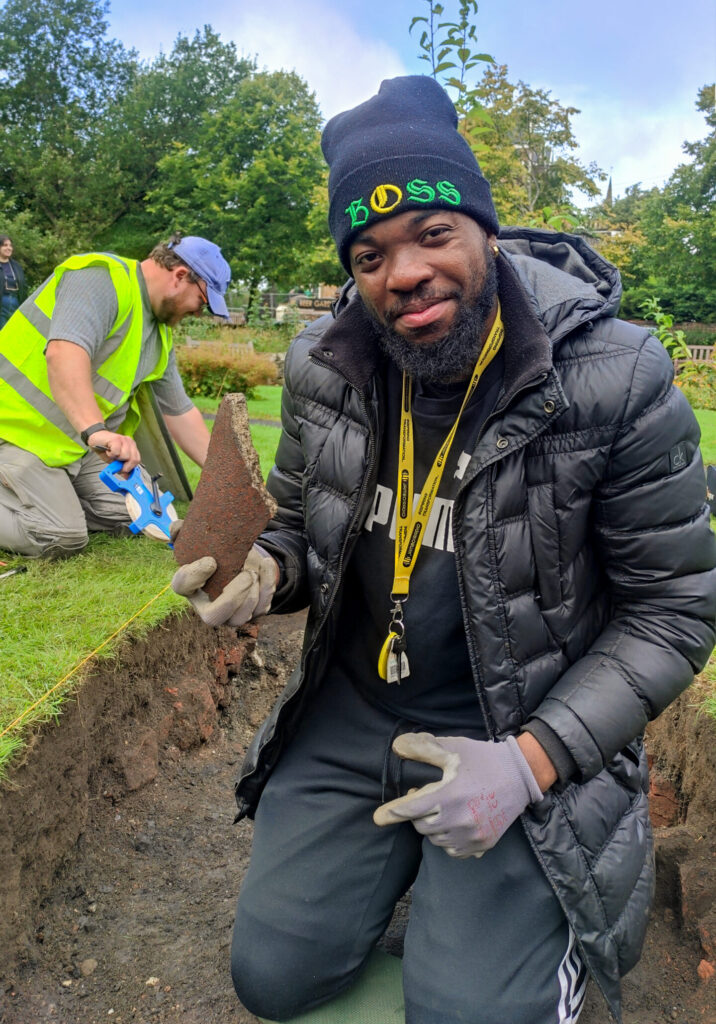 Photo of a man in a hat and black jacket in an archaeological trench holding a piece of clay pipe