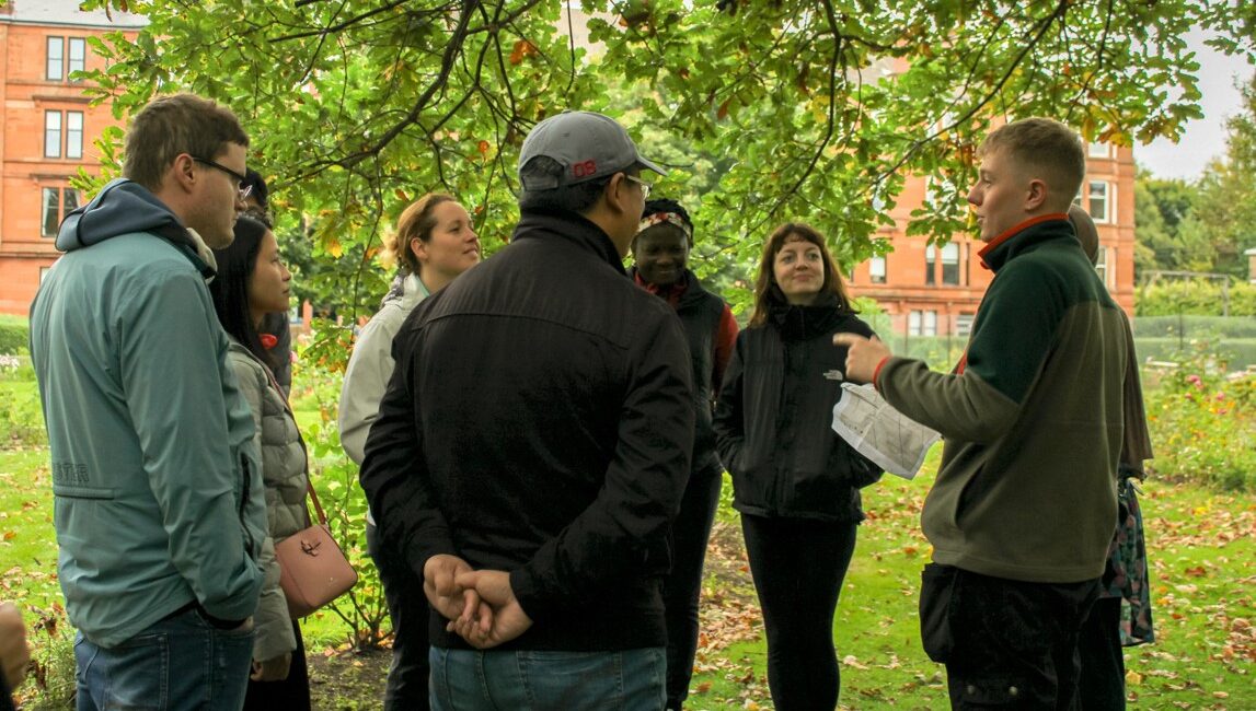People standing in a group under a tree in an urban setting listening to someone talk