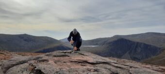 Photo of a person in warm outdoor clothing taking a sample from a large flat rock high in the mountains