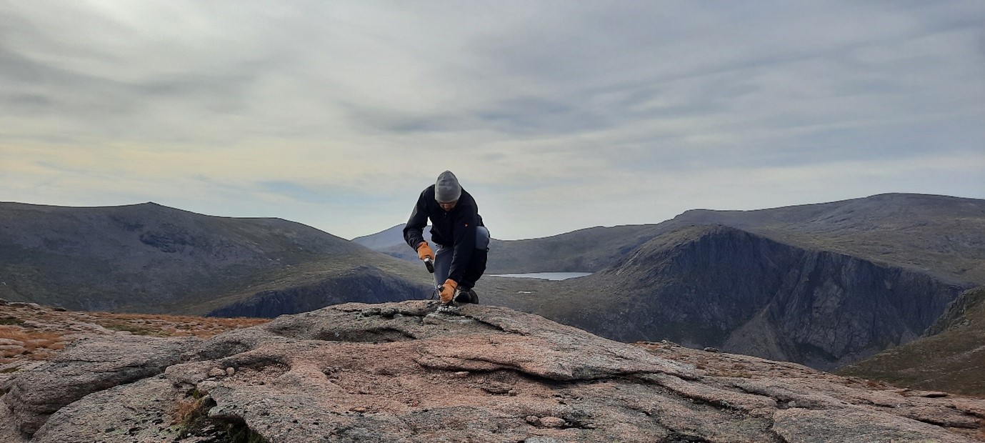 Photo of a person in warm outdoor clothing taking a sample from a large flat rock high in the mountains
