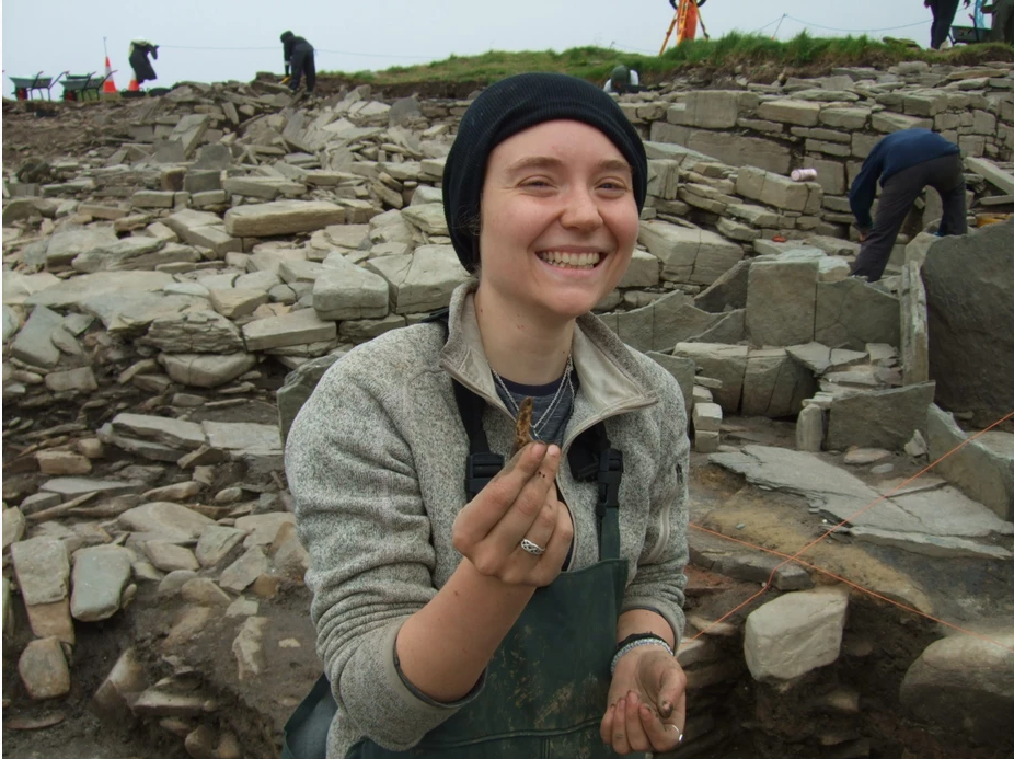 Photo of a person in a black wool hat in an archaeological trench and holding a small seal tooth