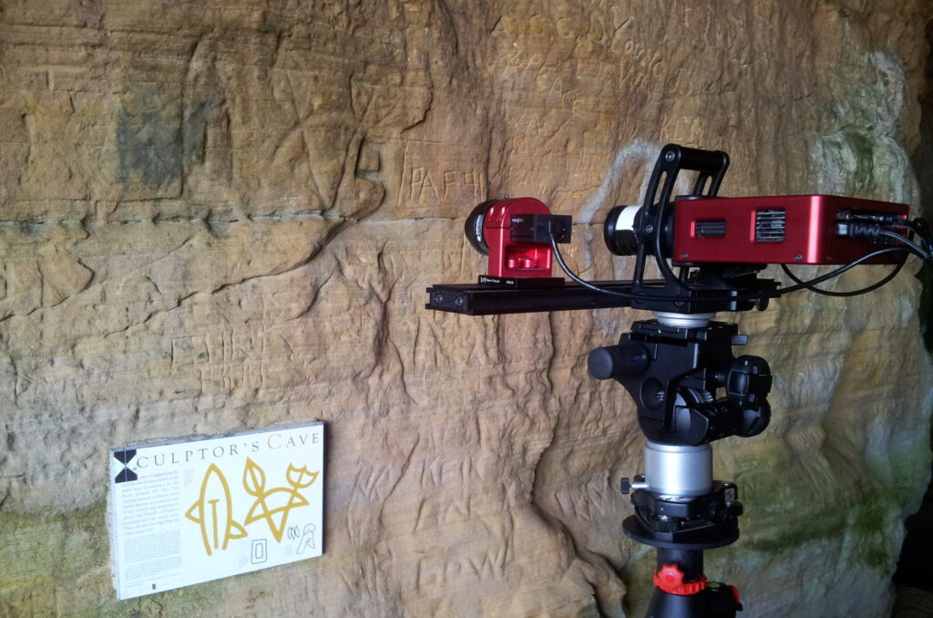 Photo of a laser scanner mounted on a tripod pointed at carvings on a cave wall