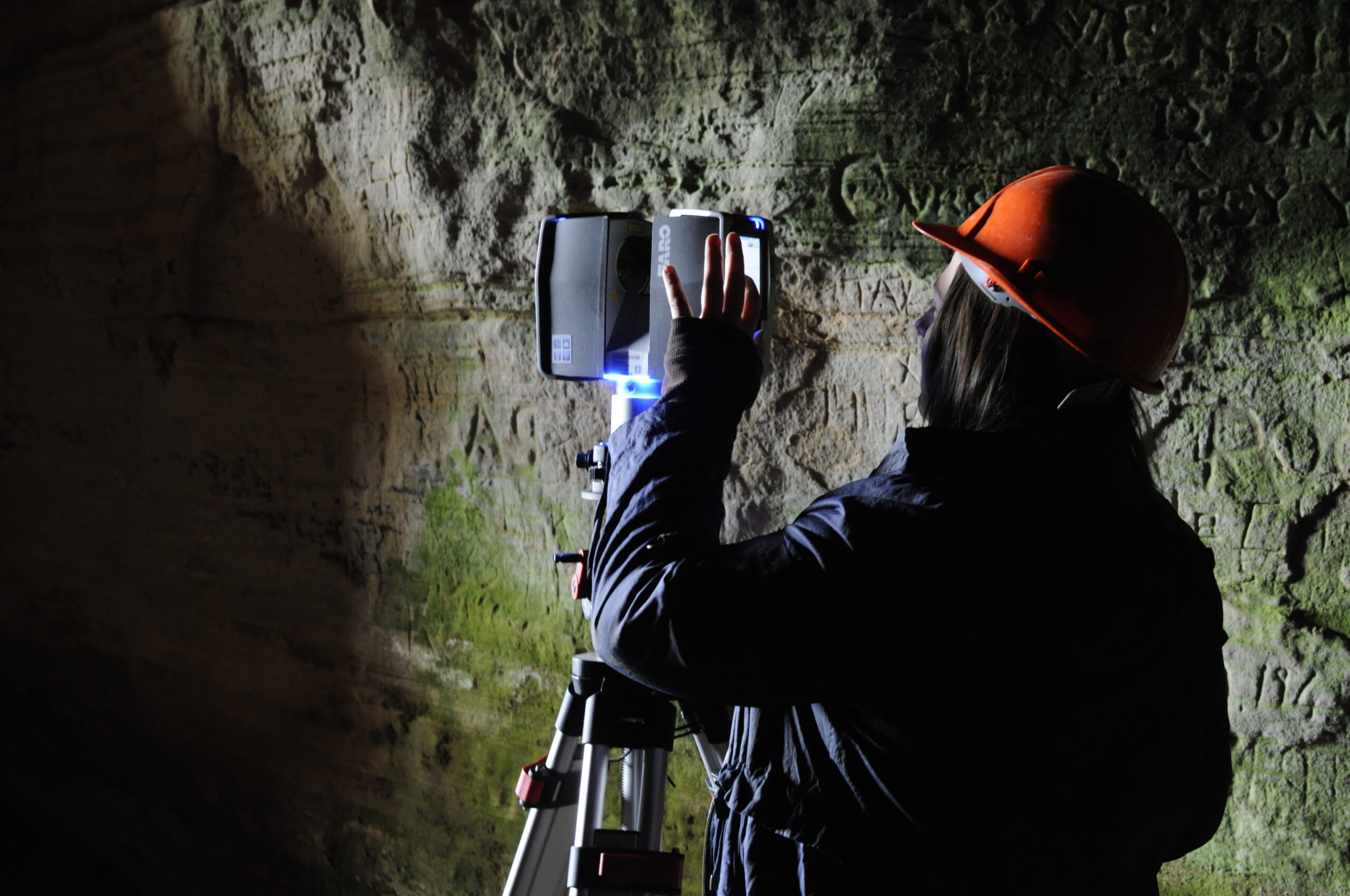 Photo of a person in an orange hard hat using a laser scanner on a tripod inside a sea cave