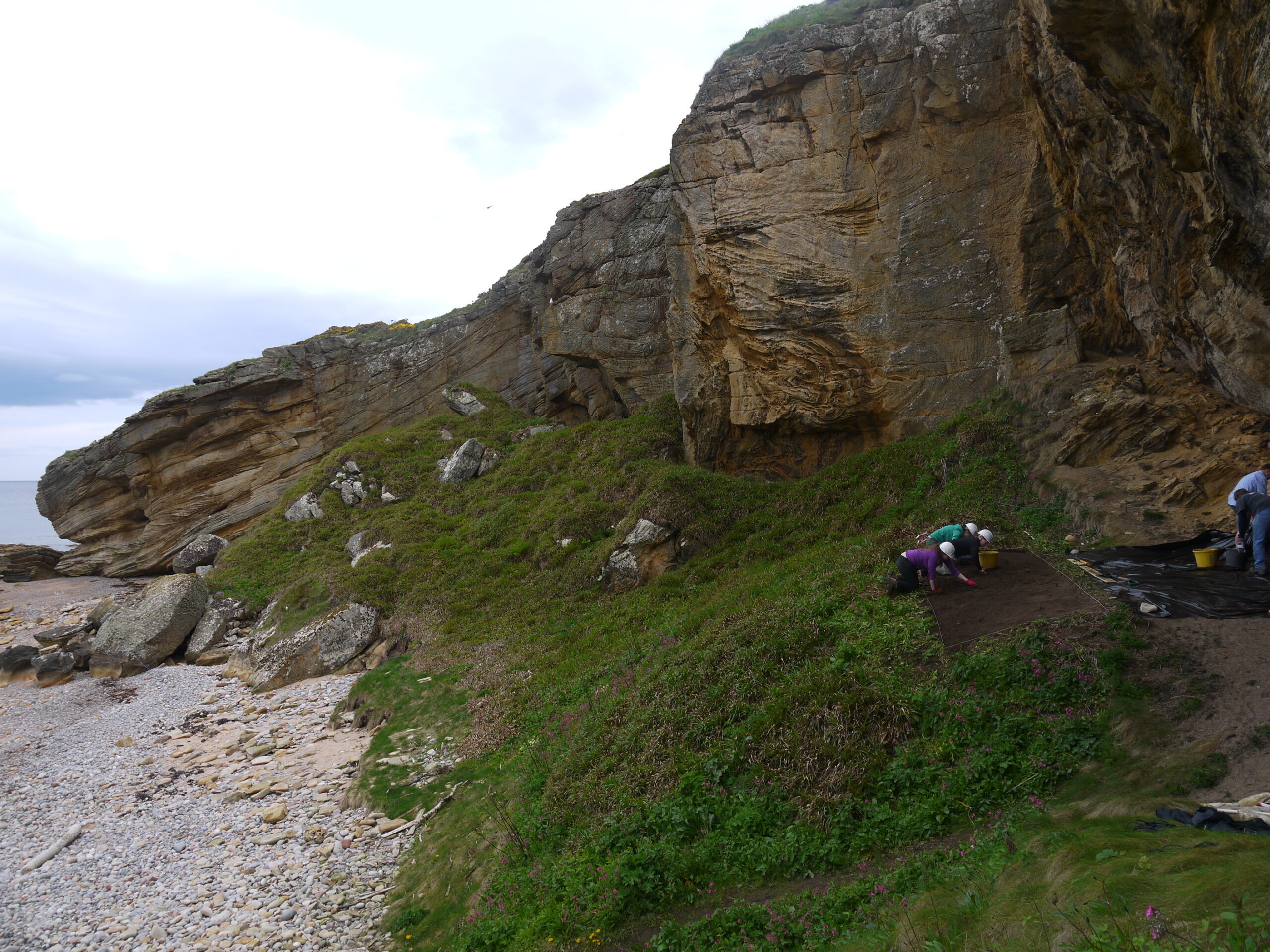 Photo of students in white hard hats kneeling and excavating a trench on a beach in front of some cliffs