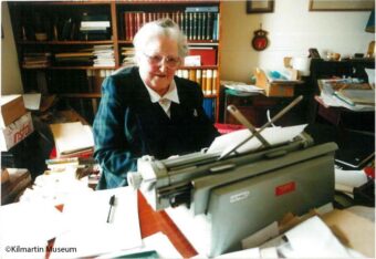 Old photo of person at a desk surrounded by papers, book, etc.