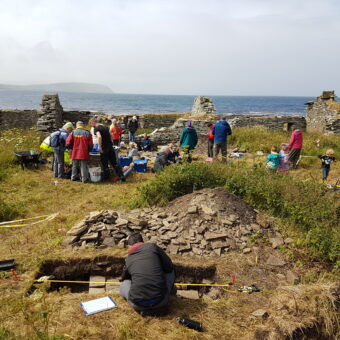 Several people standing around an archaeological site with stone ruins in the background by the water