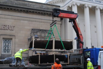 Photo of a huge wooden artefact being craned off a flat bed lorry in front of a museum