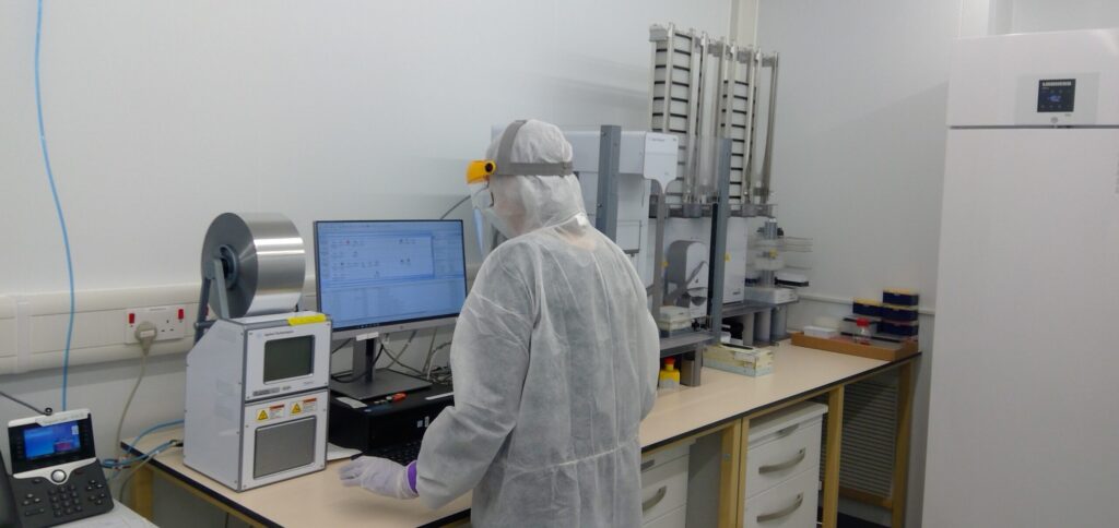 Photo of a person in white lab overalls and mask working in a lab