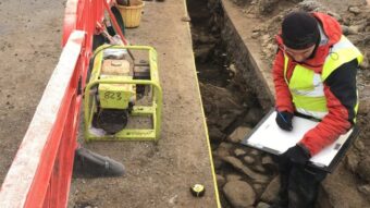Photo of a person in red jacket and high-vis vest sat in an archaeological trench drawing features