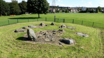 Stone circle with all but one stones laying down - behind a fence beside houses
