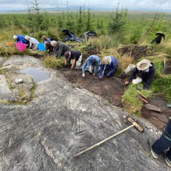 A group of people in waterproof clothing sitting or kneeling in a line cleaning a large flat rock surface covered in markings in front of them, with short pine trees and moorland in the background