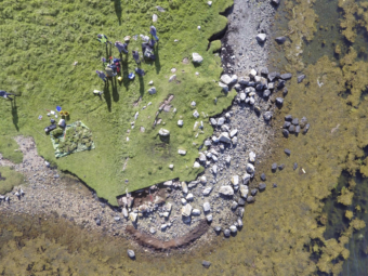 An overhead aerial image of a prehistoric site on the edge of the sea, eroding into the water, with a group of people standing next to and recording the stone remains