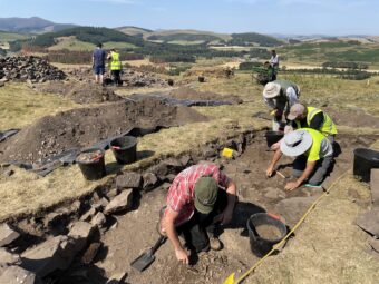 A group of people on a sunny day standing or kneeling in several archaeological trenches on the flat summit of a hill with other hills in the background