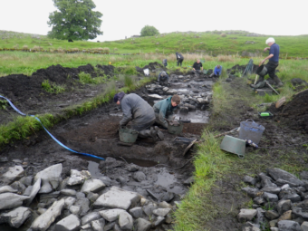 Several people in dark, waterproof clothing and wellies walking or kneeling in a long, muddy archaeological trench in a green field, with a pile of stones in the trench in the foreground and large trees in the background