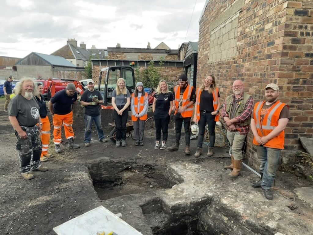 Around 12 people, many wearing orange reflective vests, standing and smiling in a semi-circle around a narrow, deep archaeological trench in an industrial area