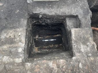 An overhead view of a small, square, deep archaeological trench cutting through stone to show the remains of a landsale coal wharf, showing the stone piers on either side of the cavity where the coal was dropped and the wooden structures underlying visible in the bottom of the trench