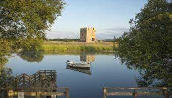 Photo of a square, stone-built tower house on an island in a river, with a boat in the foreground