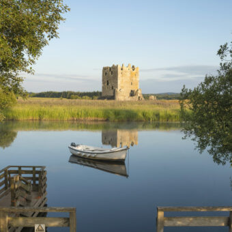Photo of a square, stone-built tower house on an island in a river, with a boat in the foreground