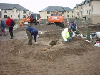 Photo of an archaeological dig in a housing estate with archaeologists revealing a stone box burial