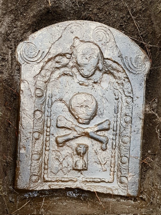 Photo of a gravestone with a skull and crossbones on it, uncovered from a trench in the ground