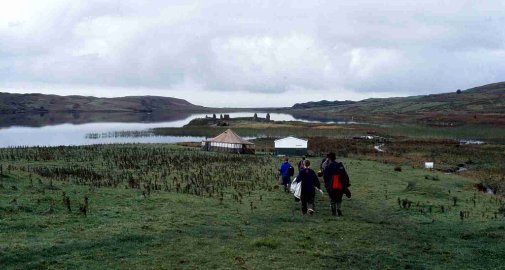 Landscape photo of a small group of people in waterproof clothing walking to a historic site, with lochs and a tent in the distance
