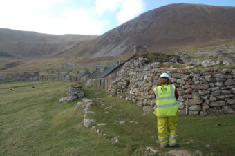 Photo of an archaeologist in a high-vis jacket and white hard hat walking next to a ruined stone settlement on an island