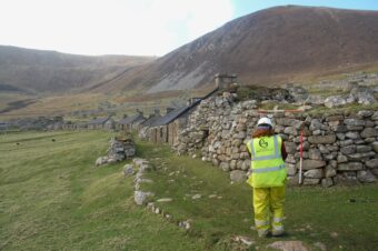 Photo of an archaeologist in a high-vis jacket and white hard hat walking next to a ruined stone settlement on an island