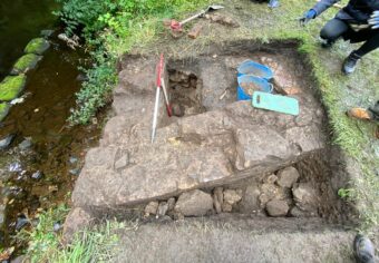 Several people standing around a trench beside a river with archaeological tools, such as a trowel and buckets, inside it.