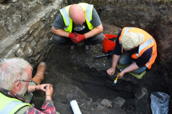 Three people wearing high-vis vests in a trench looking down