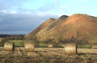 Photo of three shale bings in the background with round haybales lying in a field in the foreground