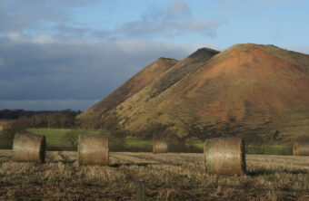 Photo of three shale bings in the background with round haybales lying in a field in the foreground