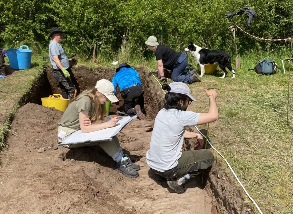Six people (some drawing, talking, etc) and a dog in and around the rectangular trench where a paved surface has been revealed 