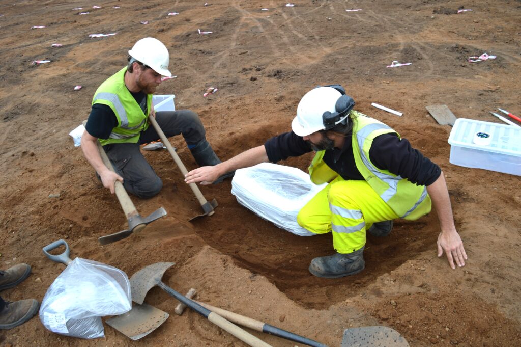 Photo of two archaeologists in white hard hats and yellow high-vis vests passing tools to each other in a small square trench
