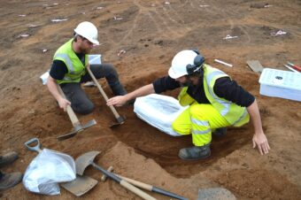 Photo of two archaeologists in white hard hats and yellow high-vis vests passing tools to each other in a small square trench