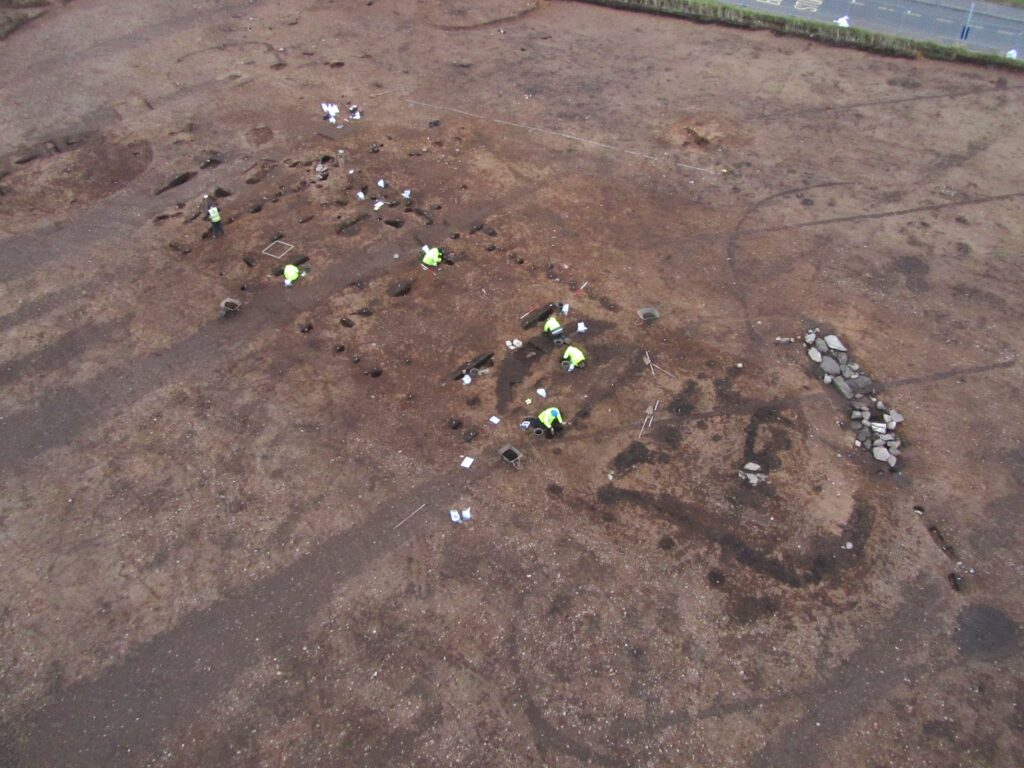 Aerial photo of a team of archaeologists in yellow high-vis jackets excavating a timber hall on a large archaeological site