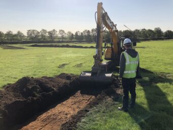 Photo of a person in white hard hat and yellow reflective vest watching a yellow and black scoop digger cutting a trench in a field