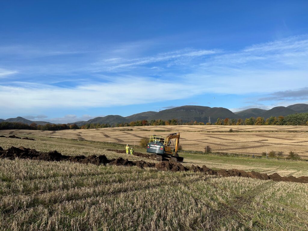 Photo of two people in white hard hats and yellow high vis clothing in an archaeological trench in a rural landscape with a yellow and black digger next to them