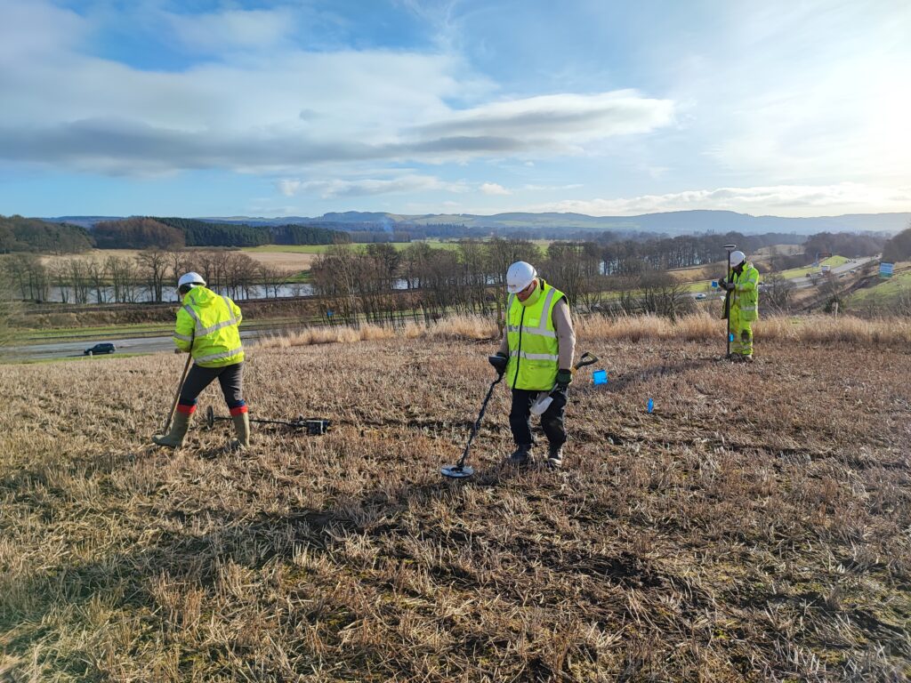 Photo of three people in yellow high-vis vests and white hard hats metal detecting in a field
