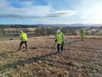 Photo of three people in yellow high-vis vests and white hard hats metal detecting in a field