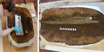 Photo of an archaeologist wearing blow plastic gloves carefully excavating a sword from a chunk of soil in a lab and