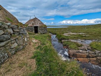 A pair of thatched buildings. The two buildings are on an oval plan, with thatched roofs weighed down with large stones. Water running beside them.