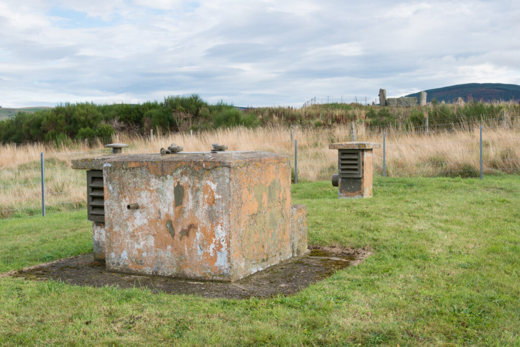Photo of a square stone military bunker in a field with a prehistoric stone circle in the background