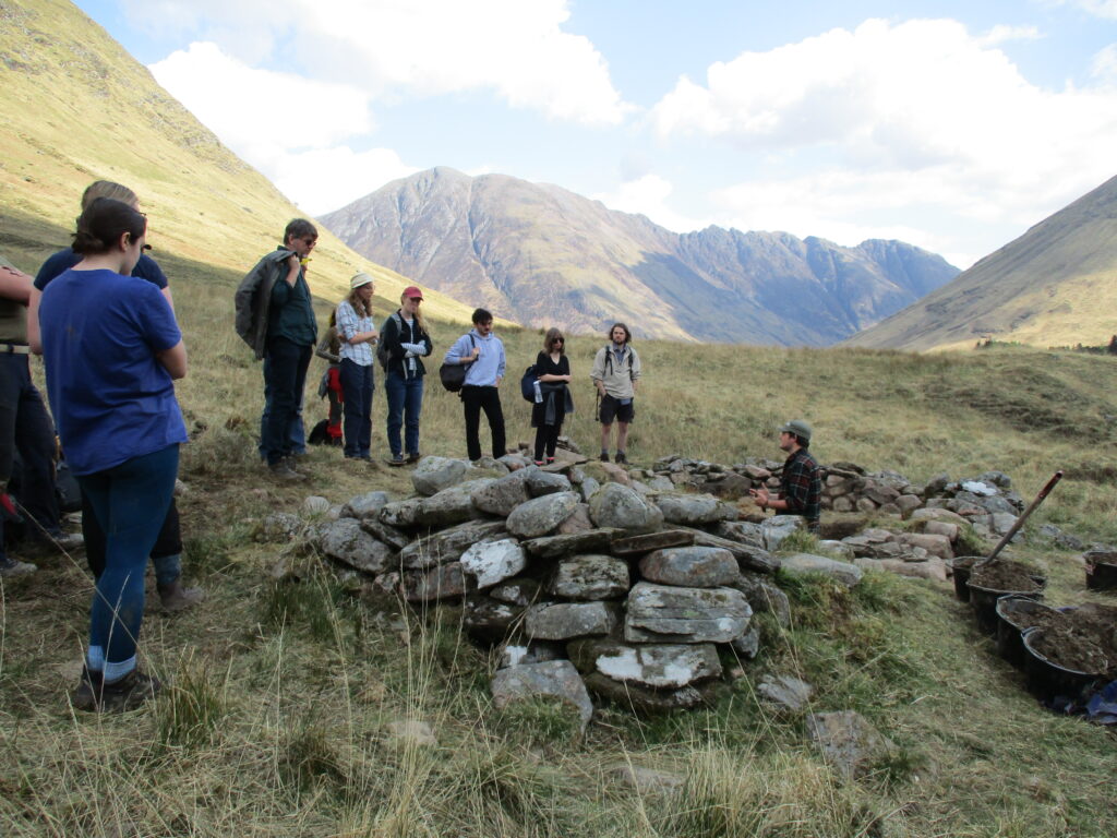 Several people listening to a person in a trench talk in a mountain setting