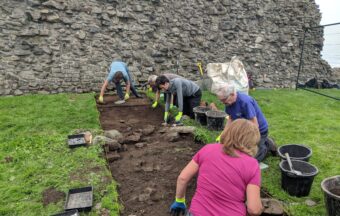 Several people digging in a trench in front of a stone castle