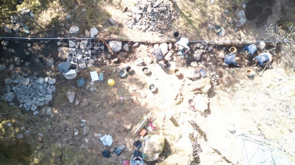 Aerial photo of an archaeological trench in a forest showing stones and buckets