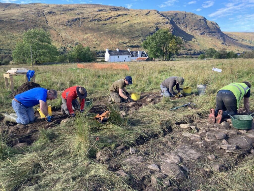 Photo of people kneeling in a line by a shallow archaeological trench in a field in the Highlands