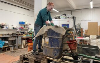 Photo of a person wearing gloves in a lab standing on wooden pallets with their hands in a large blue drum