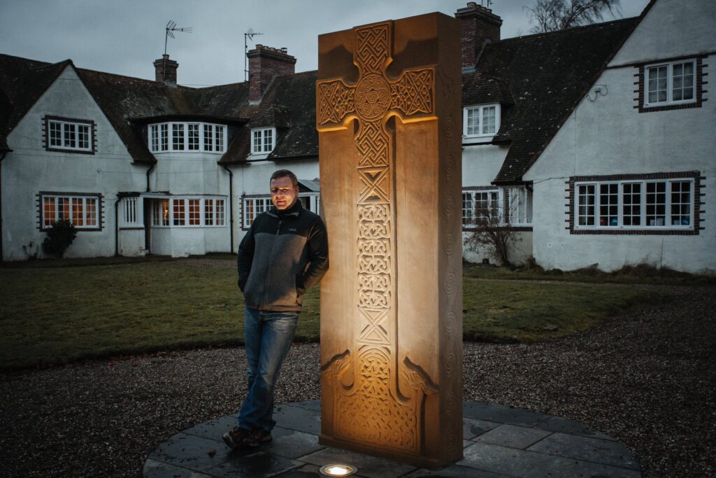 Photo of a man leaning against a large carved stone slab in front of a large white house; the slab has a large Christian cross carved into it with interlaced knotwork